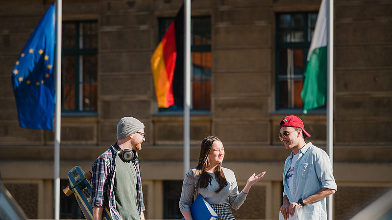 Die europäische, deutsche und sächsische Flagge fliegen vor einem Hochschulgebäude in Görlitz. Davor sind drei Studierende im Gespräch.